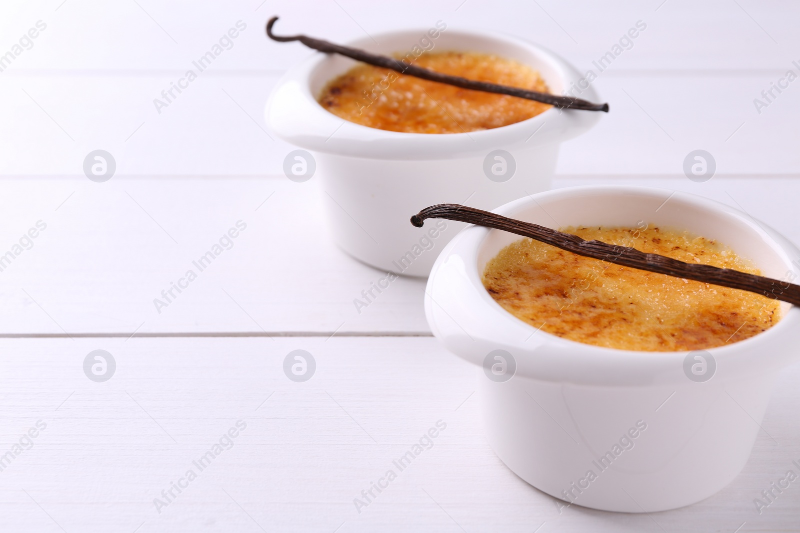 Photo of Delicious creme brulee in bowls and vanilla pods on white wooden table, closeup. Space for text