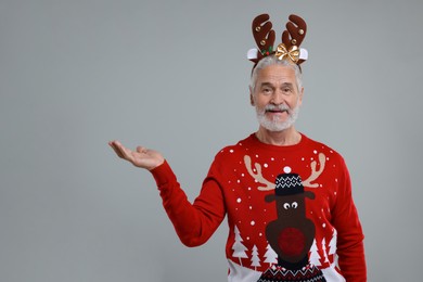 Photo of Senior man in Christmas sweater and reindeer headband showing something on grey background. Space for text