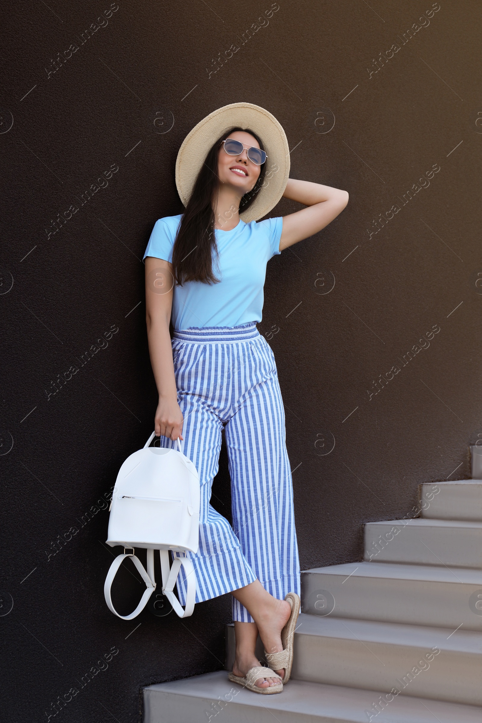 Photo of Beautiful young woman with stylish backpack and hat on stairs leaning to black wall