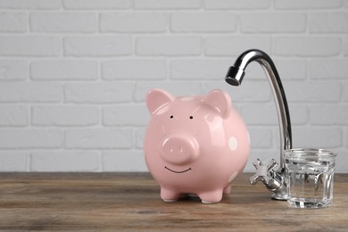 Photo of Water scarcity concept. Piggy bank, tap and glass of drink on wooden table, space for text