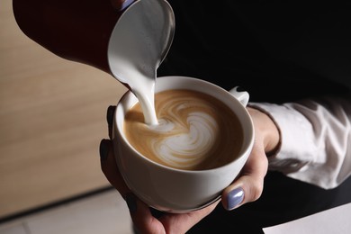 Photo of Barista pouring steamed milk from pitcher into cup of aromatic coffee in cafe, closeup