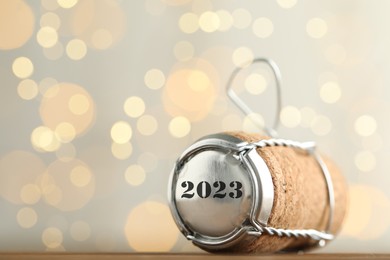 Cork of sparkling wine and muselet cap with engraving 2023 on wooden table against blurred festive lights, space for text. Bokeh effect