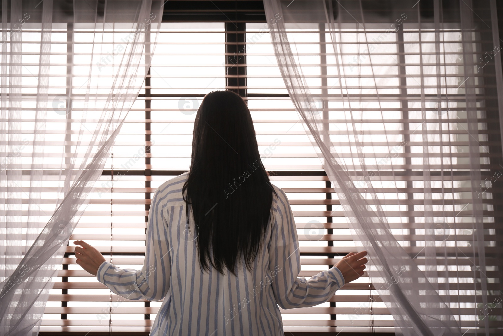 Photo of Woman opening window curtains at home in morning, back view