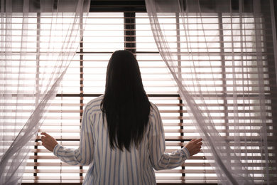 Woman opening window curtains at home in morning, back view