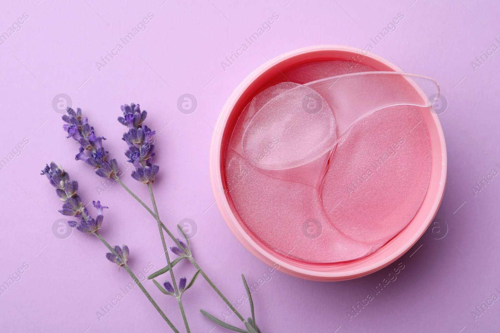 Photo of Under eye patches in jar with spatula and lavender flowers on lilac background, flat lay. Cosmetic product