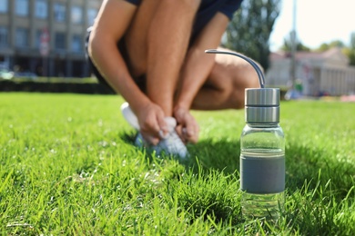 Photo of Bottle with water near man lacing his sneakers outdoors. Space for text