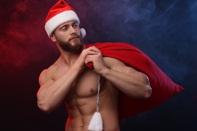 Photo of Muscular young man in Santa hat holding bag with presents on color background