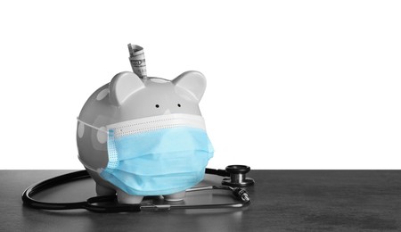Photo of Piggy bank in protective mask and stethoscope on dark grey table against white background, space for text. Medical insurance