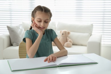 Photo of Cute little girl reading book at desk in room. Space for text