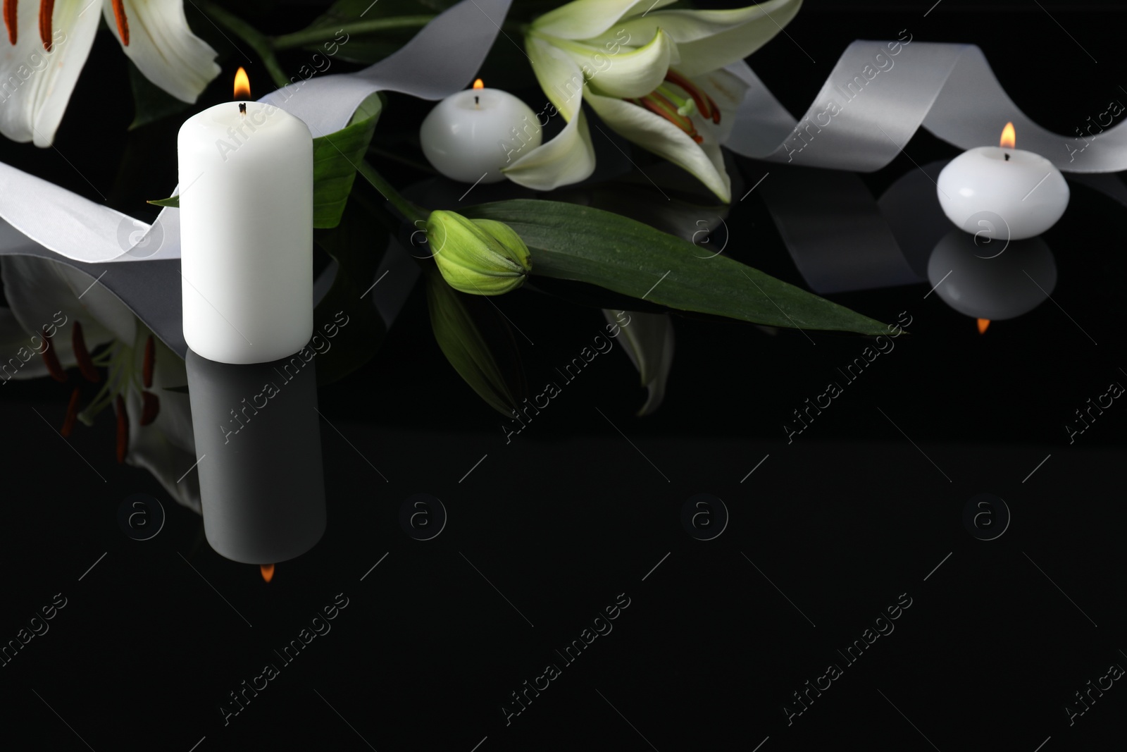 Photo of White lilies and burning candles on black mirror surface in darkness, space for text. Funeral symbols