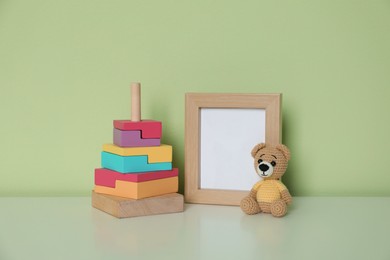 Photo of Empty photo frame, toy bear and pyramid on white table near light green wall. Space for design