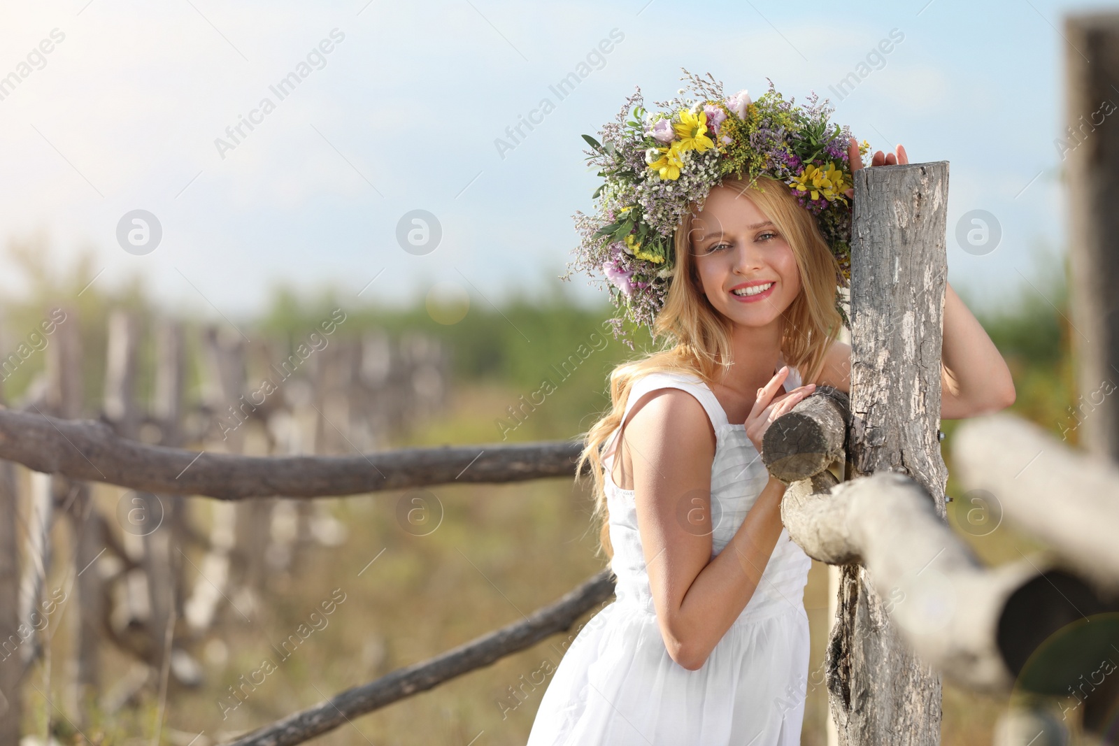 Photo of Young woman wearing wreath made of beautiful flowers near wooden fence on sunny day