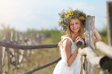 Photo of Young woman wearing wreath made of beautiful flowers near wooden fence on sunny day