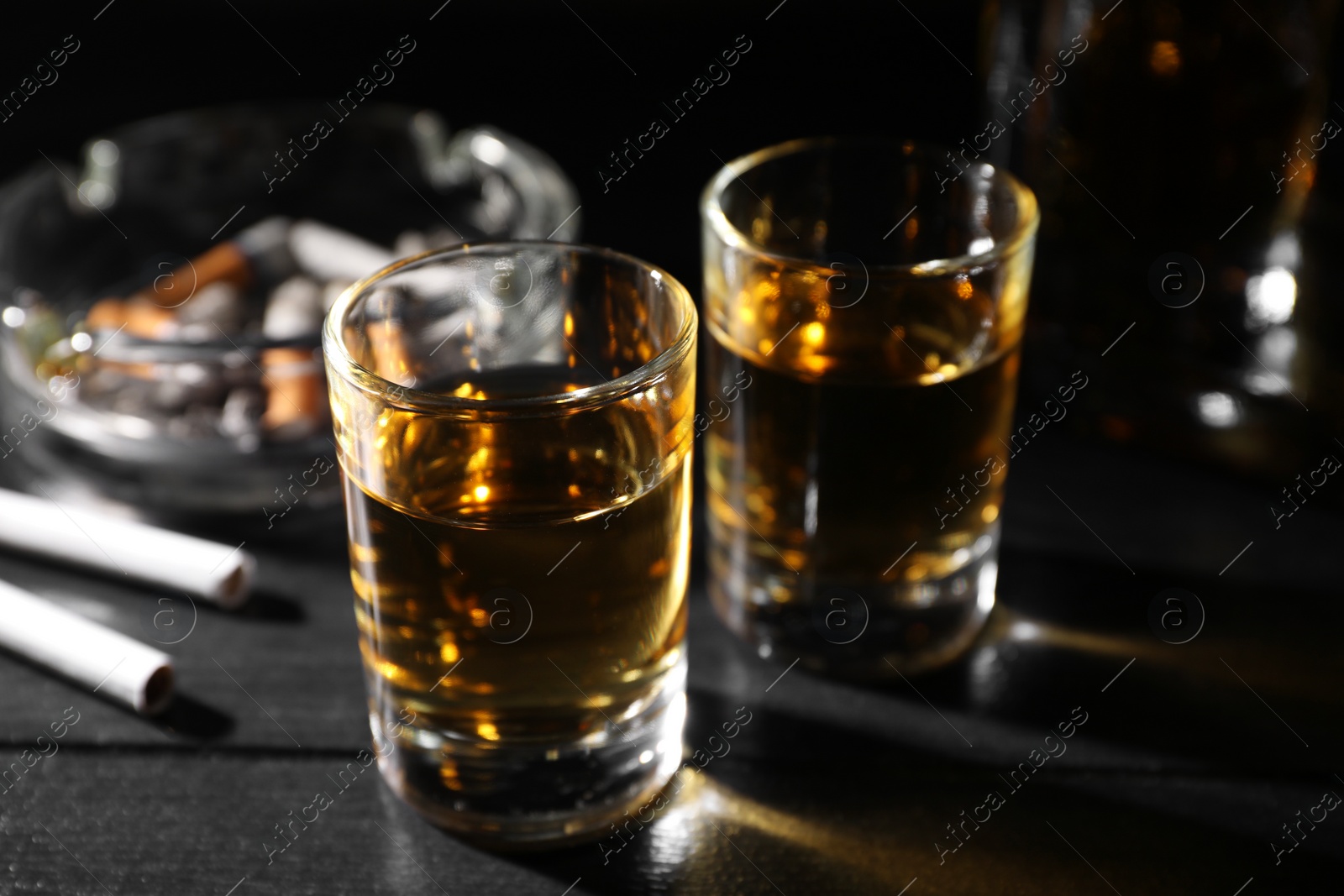 Photo of Alcohol addiction. Whiskey in glasses, cigarettes and ashtray on black wooden table, closeup