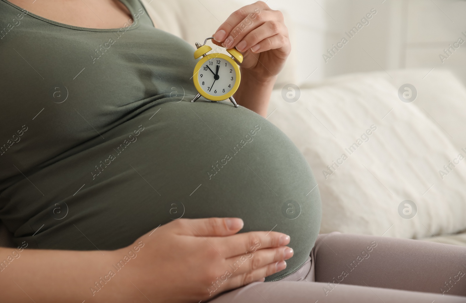 Photo of Young pregnant woman holding alarm clock near her belly at home, closeup. Time to give birth