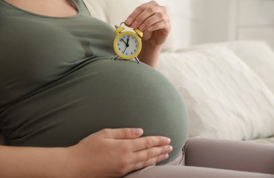 Young pregnant woman holding alarm clock near her belly at home, closeup. Time to give birth