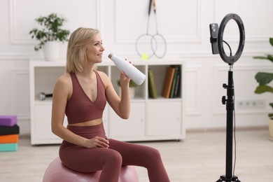 Photo of Smiling sports blogger holding bottle while streaming online fitness lesson at home