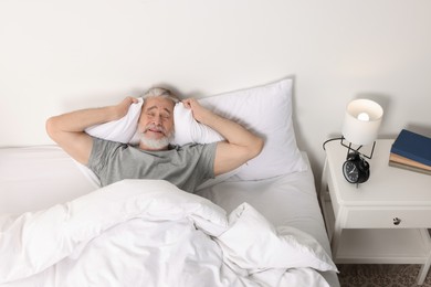 Photo of Senior man covering ears with pillows in bed and alarm clock on bedside table at home