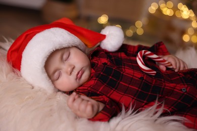 Photo of Cute baby in Santa hat with candy cane sleeping on soft faux fur indoors. Christmas season