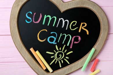 Heart shaped blackboard with text SUMMER CAMP, drawing and chalk sticks on wooden background, flat lay