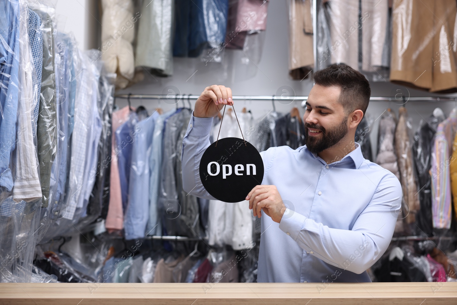 Photo of Dry-cleaning service. Happy worker holding Open sign at counter indoors, space for text