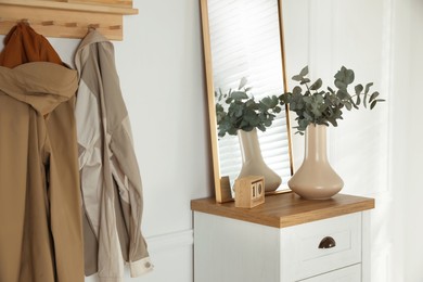 Photo of Vase with beautiful eucalyptus branches, wooden block calendar and mirror on chest of drawers near coat rack in hallway