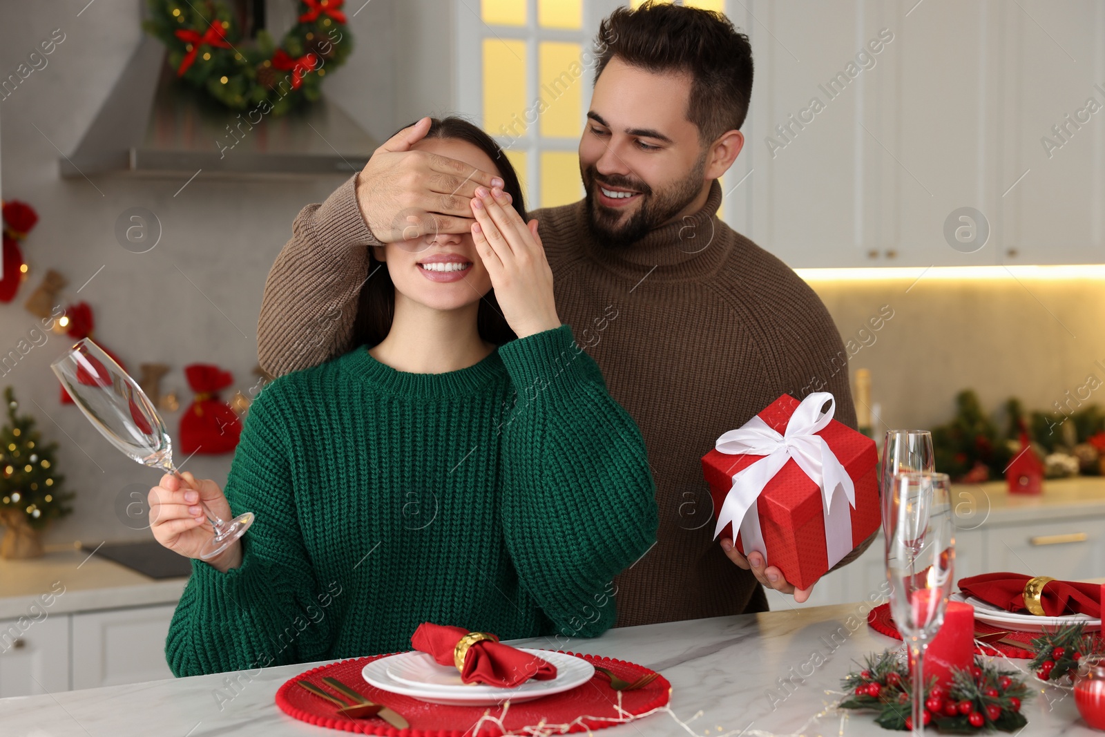 Photo of Happy young man surprising his girlfriend with Christmas gift at table in kitchen