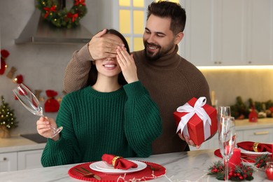 Photo of Happy young man surprising his girlfriend with Christmas gift at table in kitchen