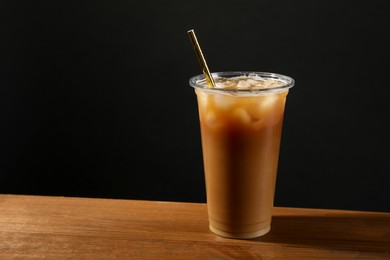 Photo of Refreshing iced coffee with milk in takeaway cup on wooden table against black background, space for text