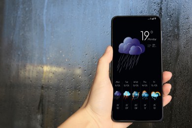 Woman checking weather using app on smartphone near wet window, closeup. Data, cloud with rain and other illustrations on screen