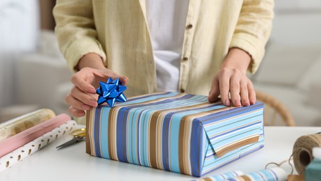 Photo of Woman wrapping gift at white table indoors, closeup