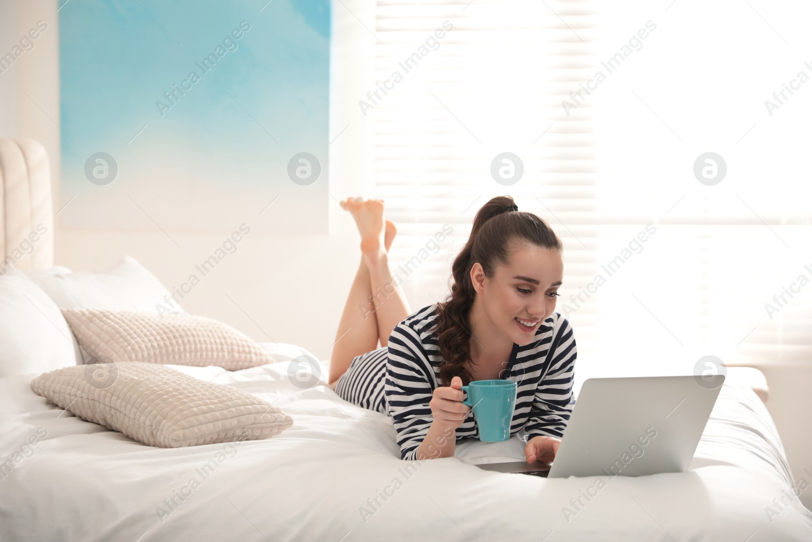 Photo of Young woman with cup of coffee and laptop on bed at home