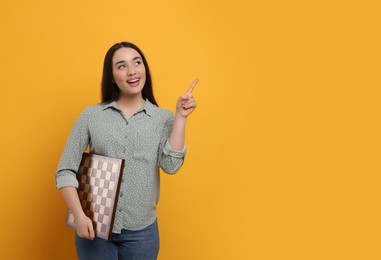 Photo of Happy woman with chessboard pointing upwards on orange background, space for text