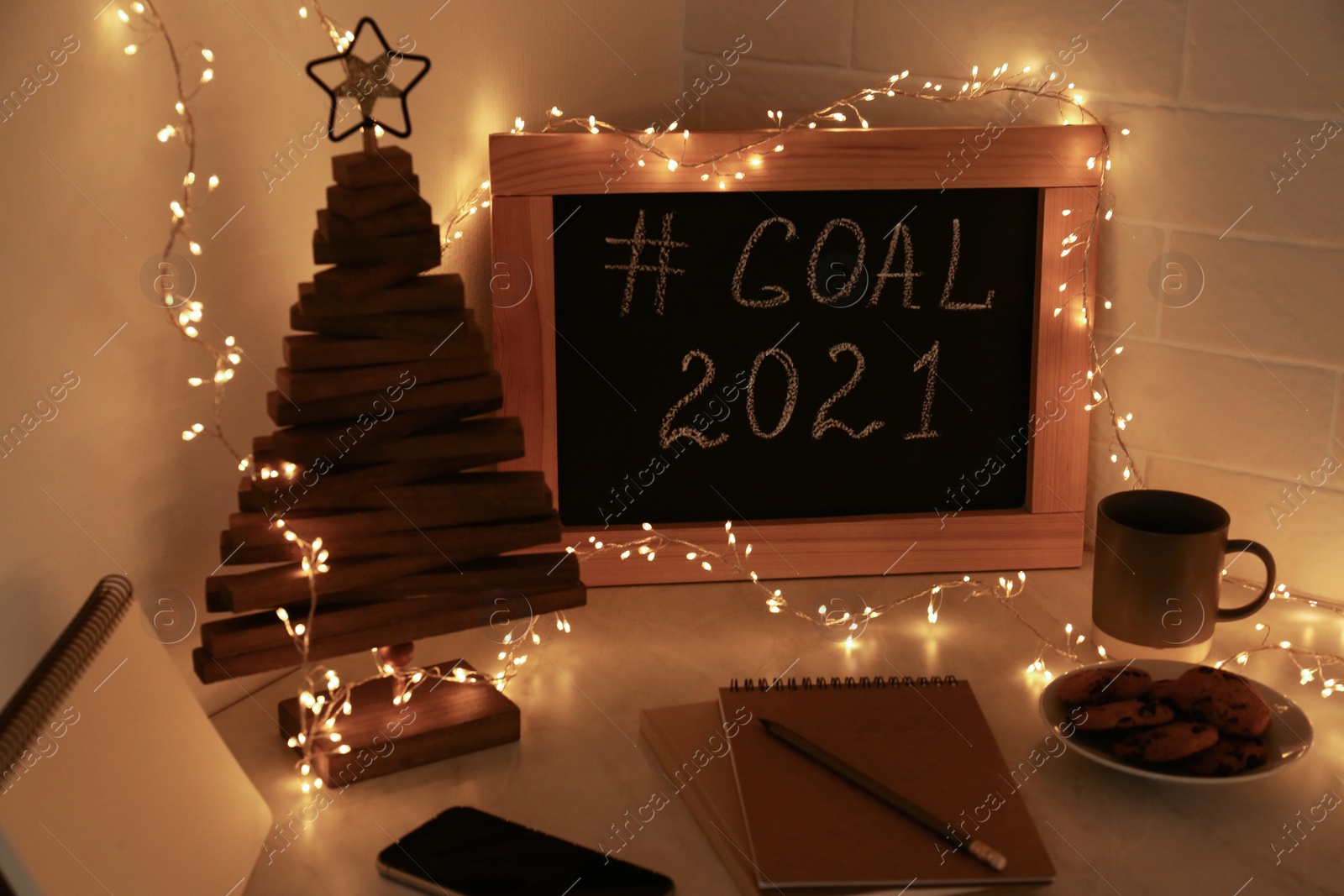 Photo of Chalkboard with hashtag Goal 2021, fairy lights near notebooks, smartphone and cookies on white table. New year targets