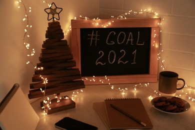 Photo of Chalkboard with hashtag Goal 2021, fairy lights near notebooks, smartphone and cookies on white table. New year targets