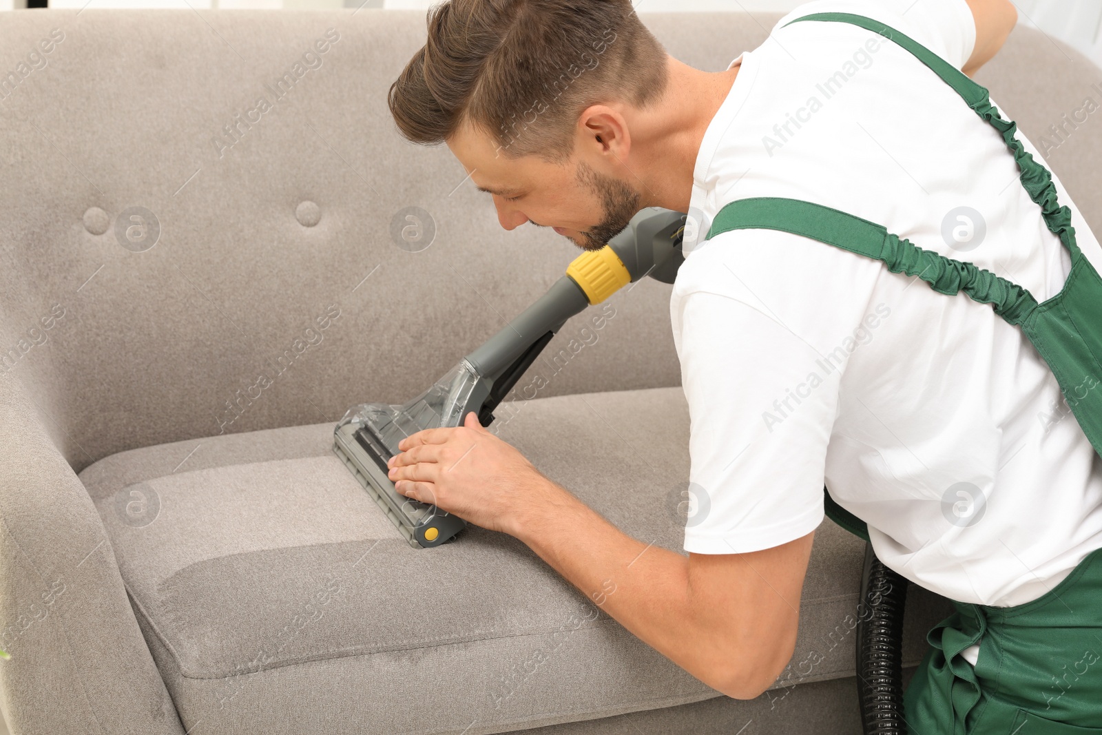Photo of Male janitor removing dirt from sofa with upholstery cleaner indoors