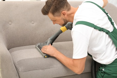 Photo of Male janitor removing dirt from sofa with upholstery cleaner indoors