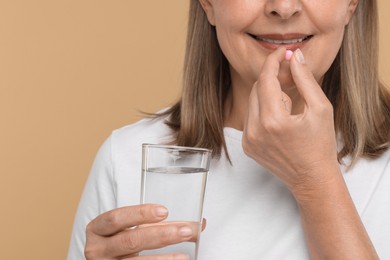 Senior woman with glass of water taking pill on beige background, closeup