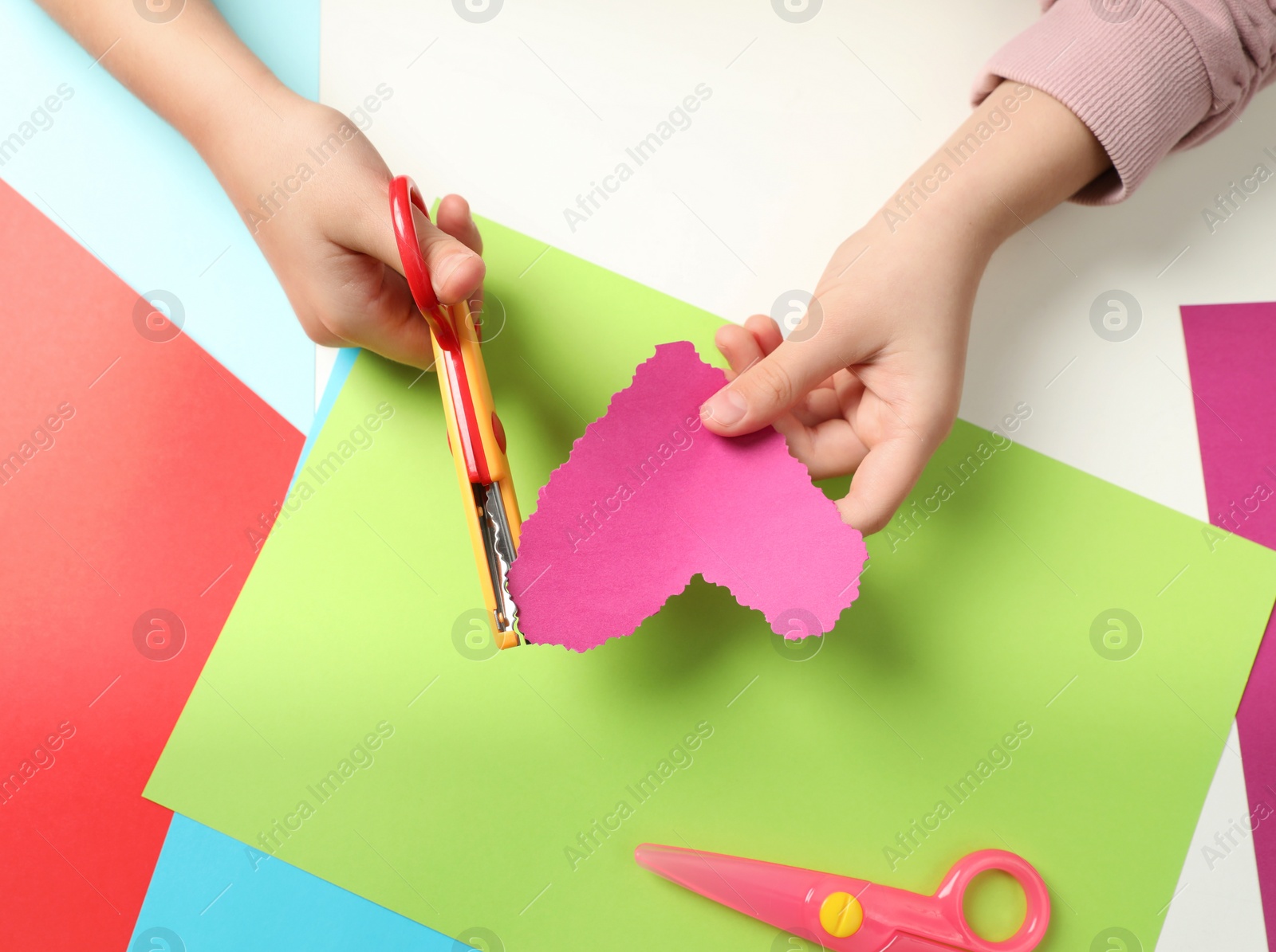 Photo of Child cutting out paper heart with craft scissors at table, top view