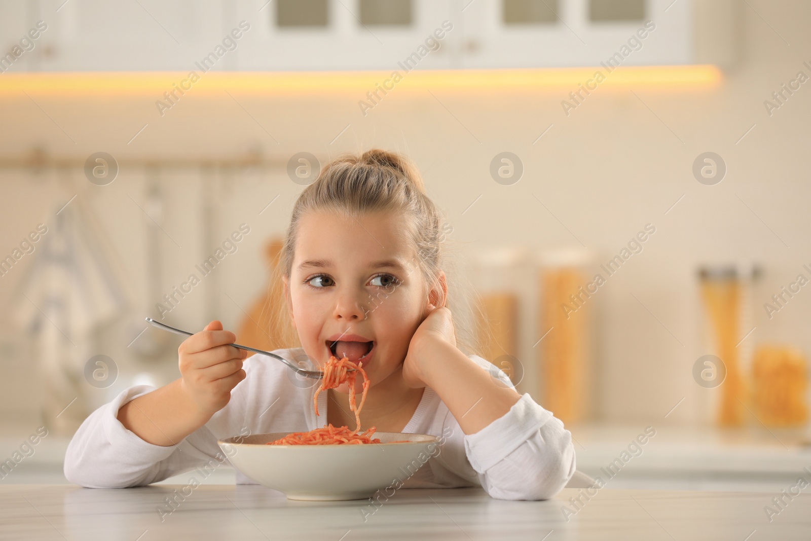 Photo of Cute little girl eating tasty pasta at table in kitchen