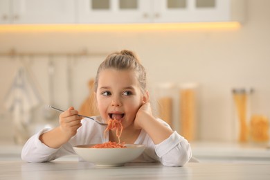 Cute little girl eating tasty pasta at table in kitchen