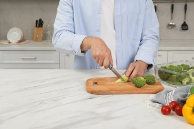 Photo of Man cutting fresh broccoli with knife near containers at white marble table in kitchen, closeup and space for text. Food storage