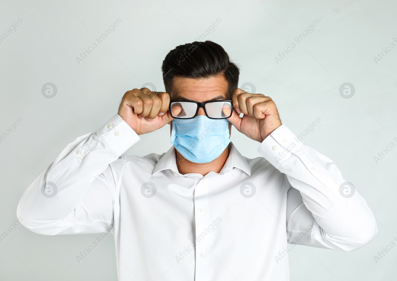 Photo of Man wiping foggy glasses caused by wearing medical mask on light background