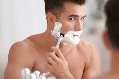 Young man shaving near mirror in bathroom