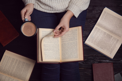 Woman with cup of coffee reading book at home, top view