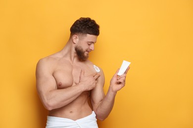 Handsome man applying moisturizing cream onto his shoulder on orange background, space for text