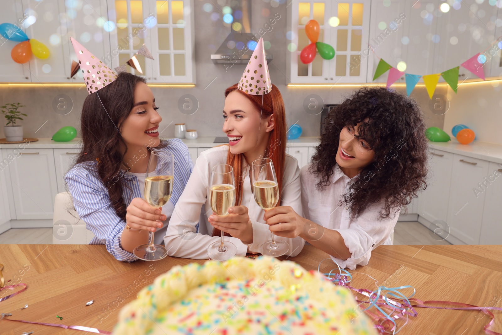 Photo of Happy young women with tasty cake and glasses of sparkling wine celebrating birthday in kitchen