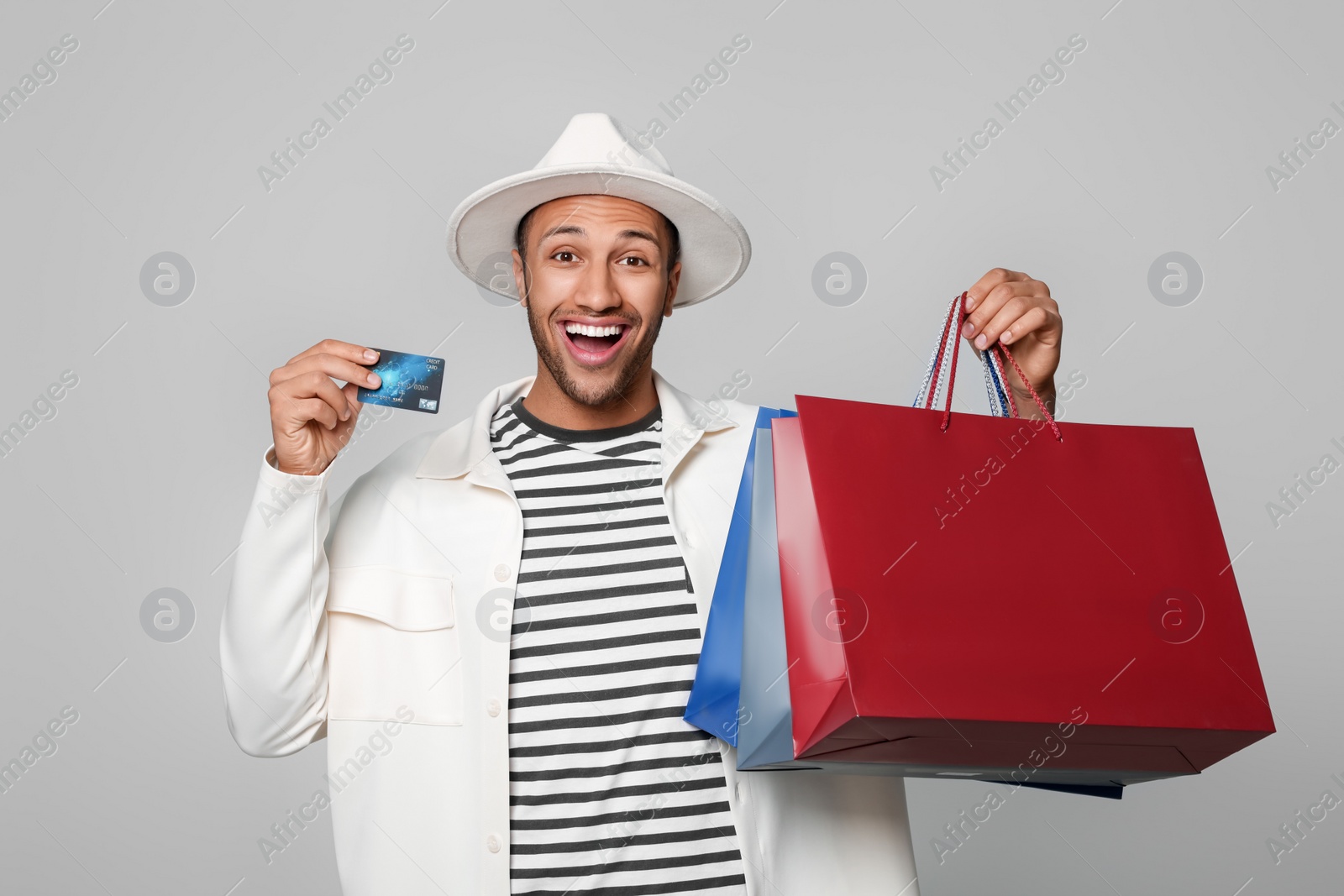 Photo of Happy African American man in hat with shopping bags and credit card on light grey background