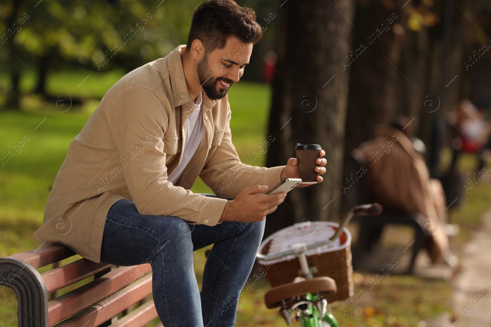 Photo of Young man sitting on bench with coffee and using smartphone in park, space for text