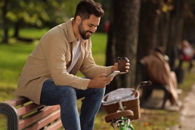 Photo of Young man sitting on bench with coffee and using smartphone in park, space for text